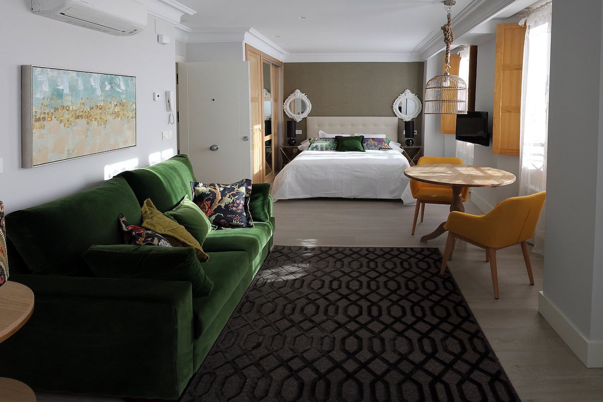 Una habitación de hotel moderna con un sofá verde, una cama con ropa blanca, una mesa de comedor con una silla amarilla y una alfombra estampada.