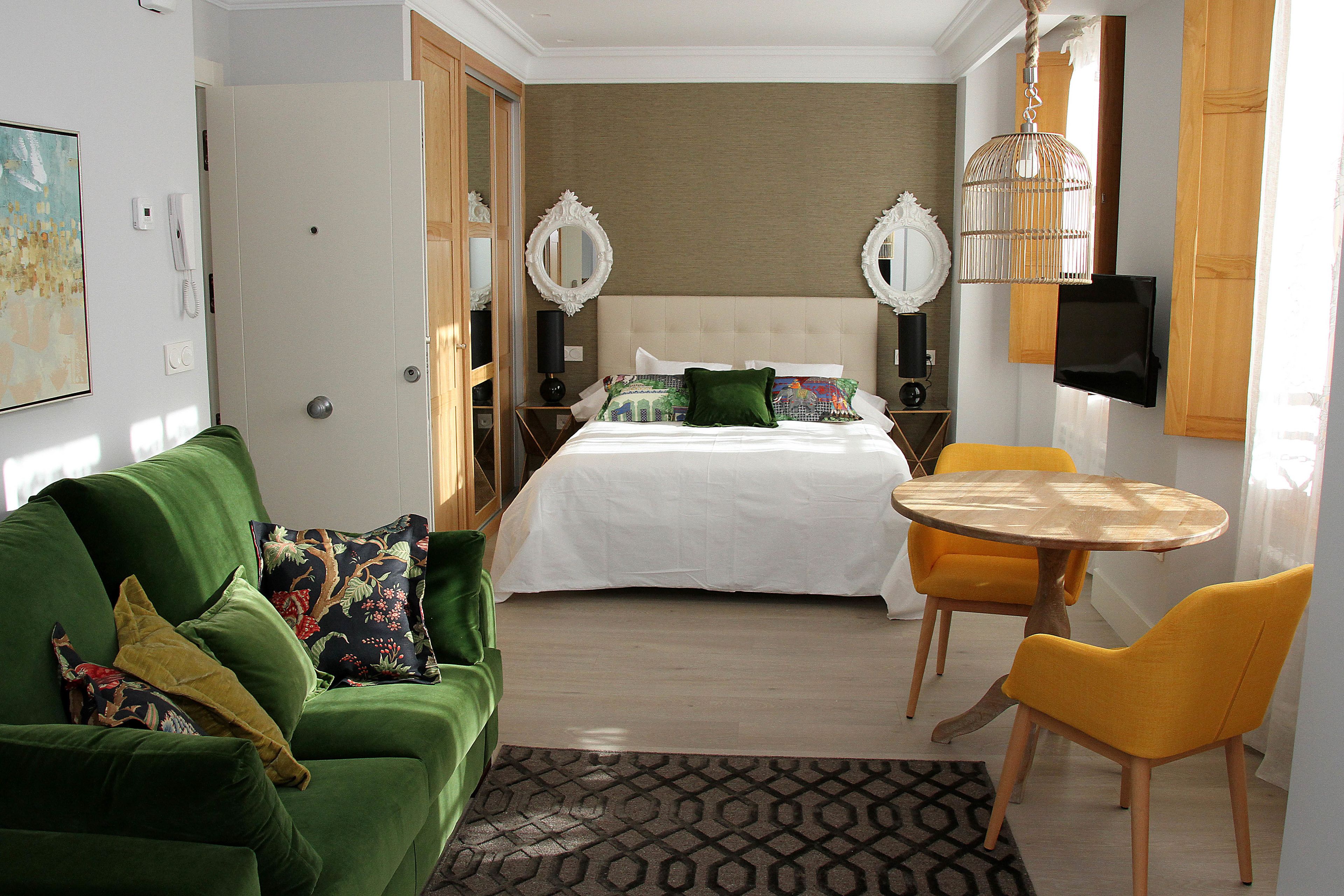 A hotel room with a bed, green sofa, yellow chairs, a small table, and a TV mounted on the wall.