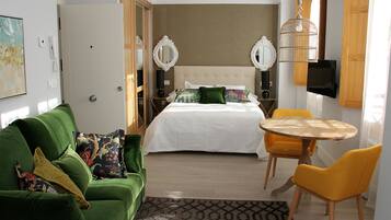 Habitación de hotel con cama, sofá verde, sillas amarillas, una mesita y un televisor montado en la pared.