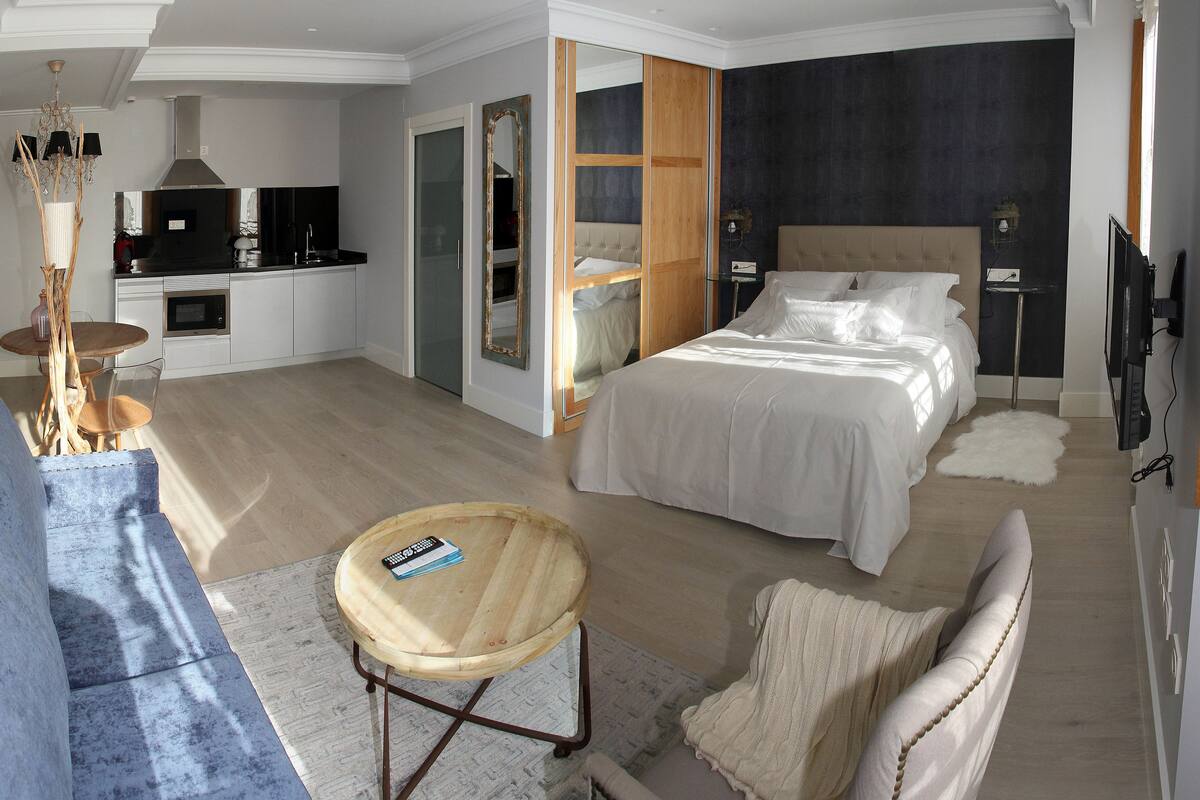 Una habitación de hotel moderna con sofá, una mesa redonda de madera, cama, televisor y un comedor con mesa y sillas.