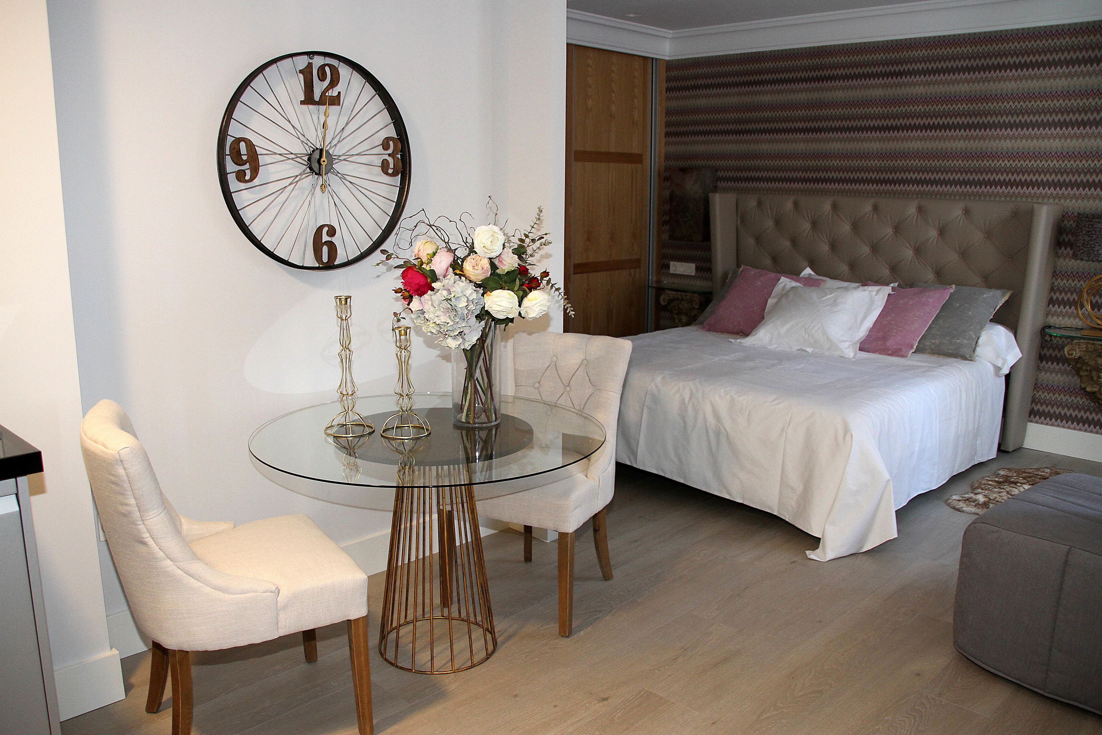 Une chambre à coucher avec un lit, une horloge murale ronde, une table en verre avec des chaises et un vase rempli de fleurs.