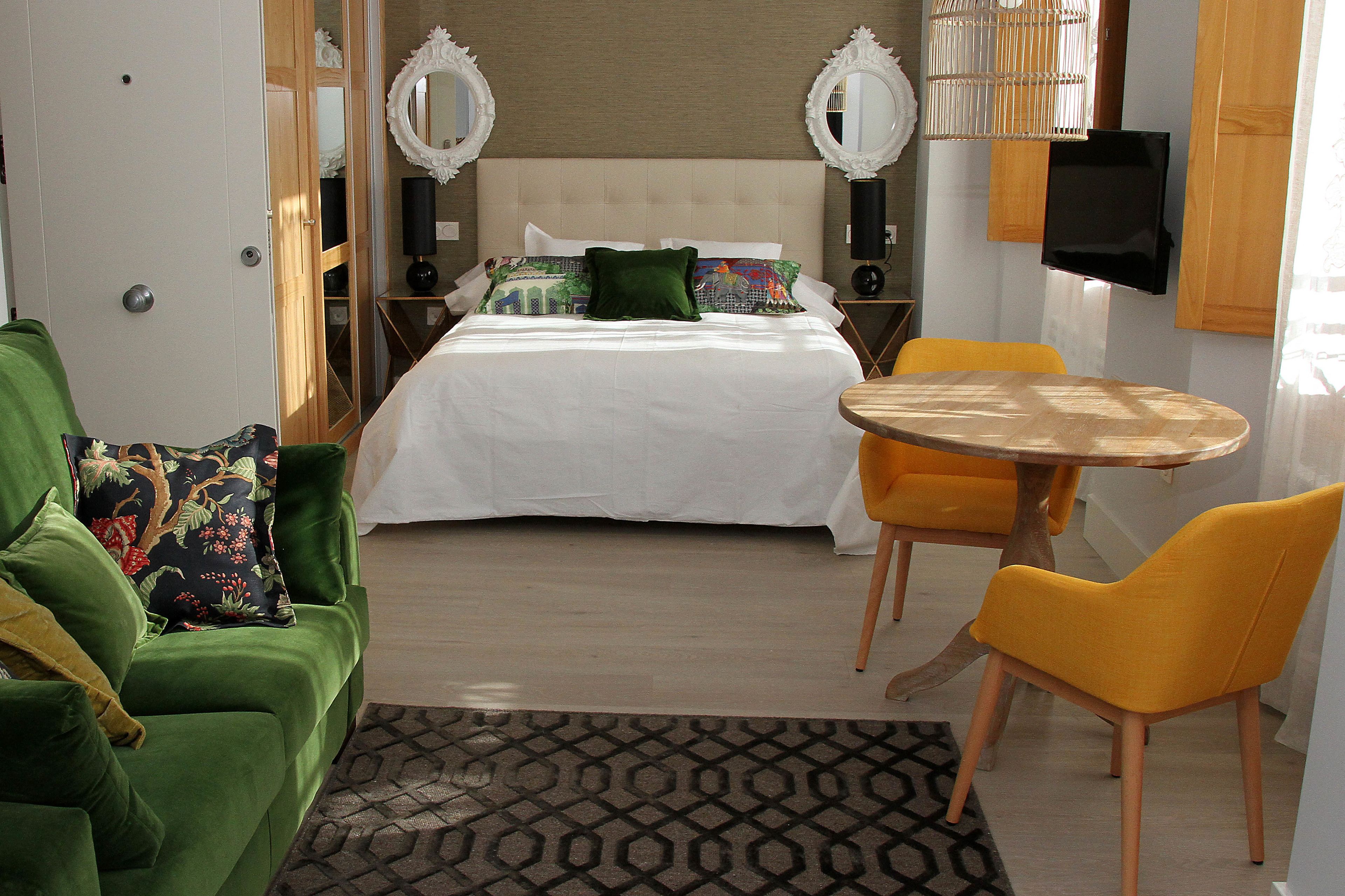 Una habitación de hotel con una cama, sillas verdes y amarillas, una mesita y un televisor.