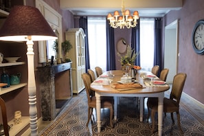 A dining room with a round table set for six, surrounded by chairs. A chandelier hangs above the table, and a clock is mounted on the wall.