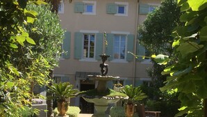 Un jardin avec une fontaine, des plantes en pot et, au fond, un bâtiment doté de volets.