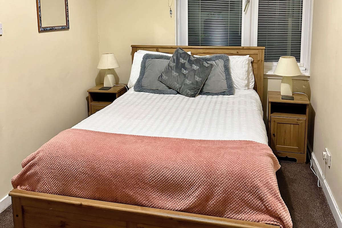 A neatly arranged bedroom with a wooden bed, two bedside tables with lamps, a window with blinds, and a wall-mounted mirror.