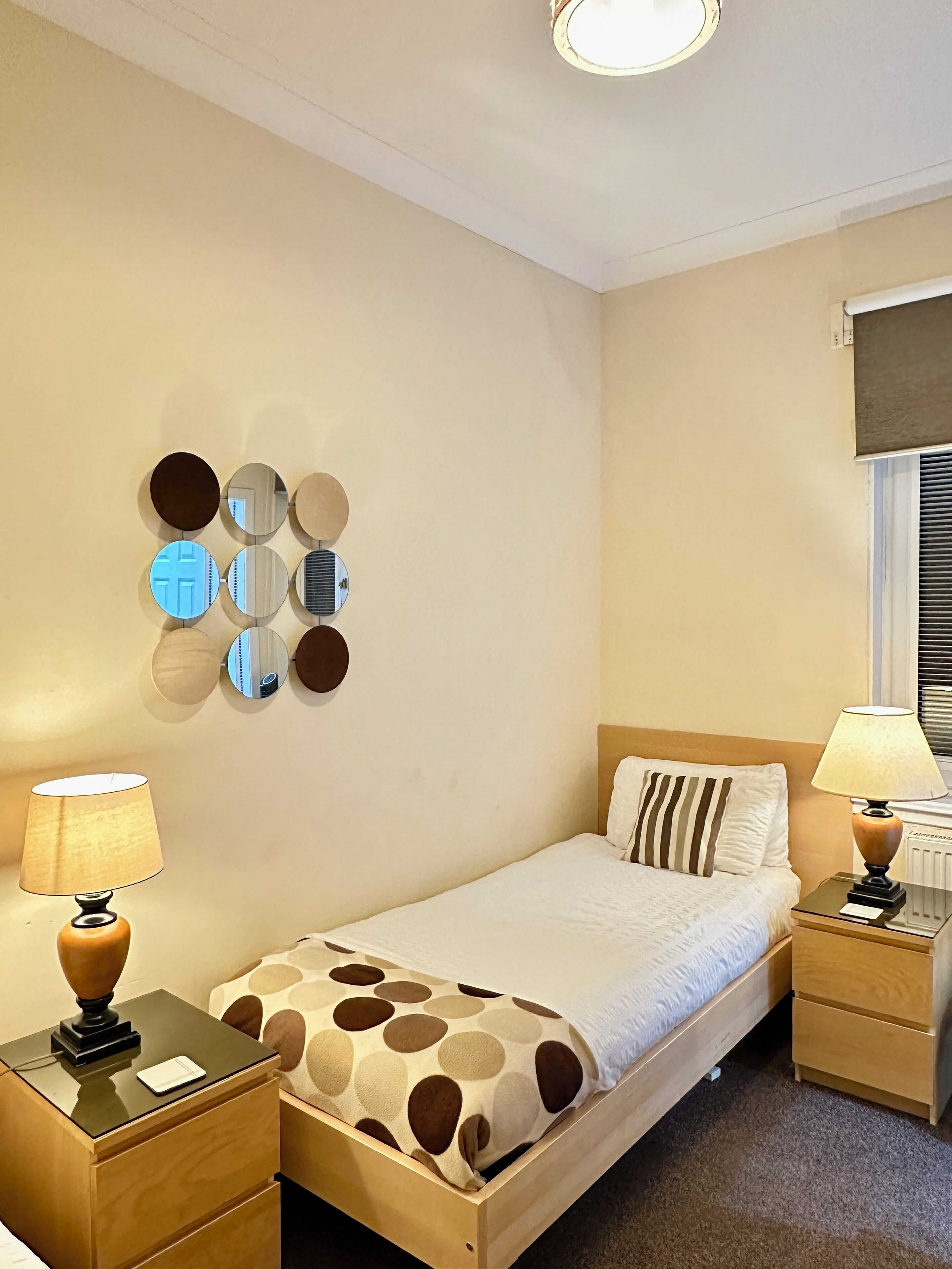 A neatly arranged bedroom with a bed, two bedside tables, a wall-mounted mirror, and a window with blinds.