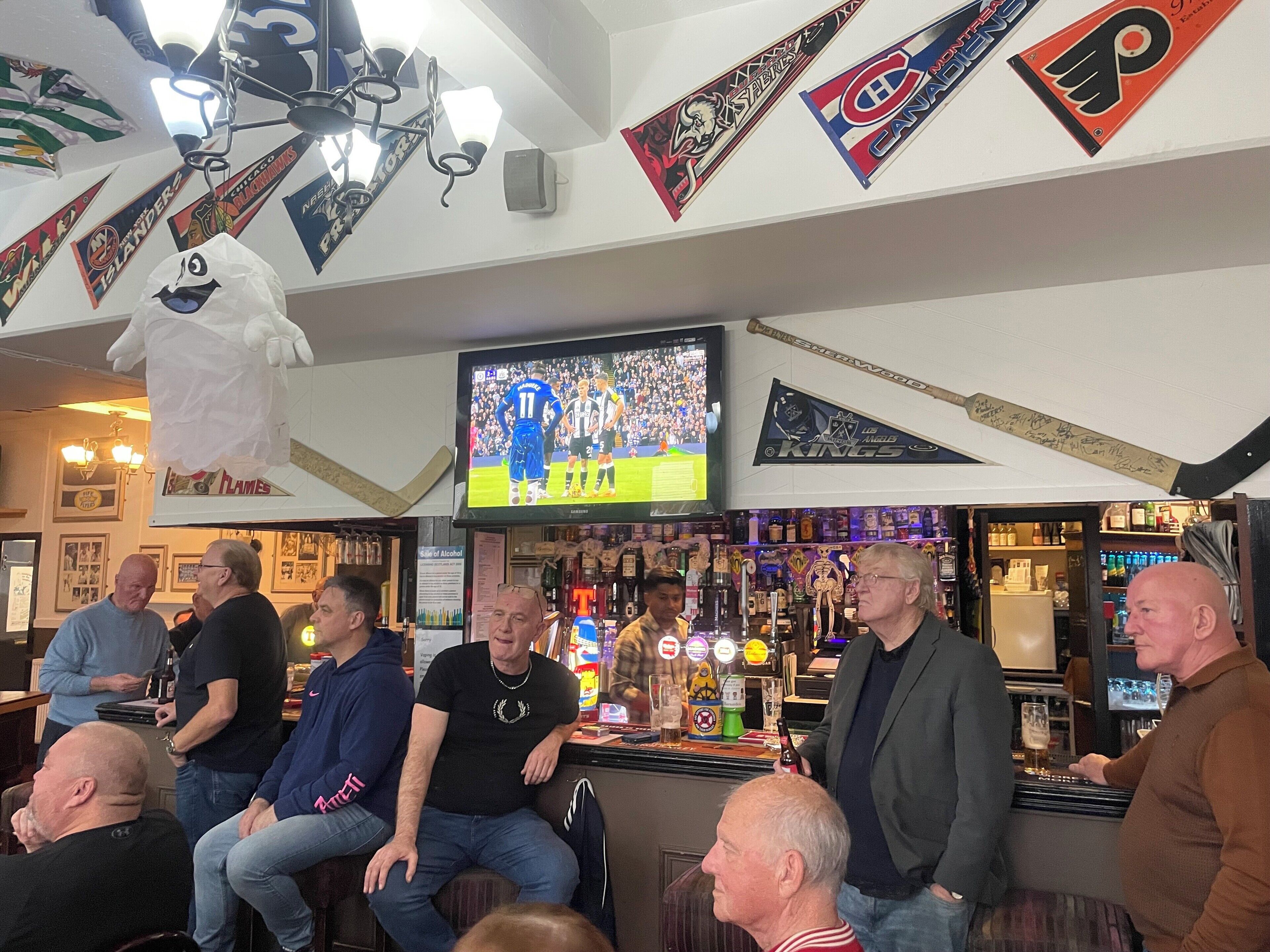 Un bar con una televisión mostrando un evento deportivo, varias banderas y un ventilador de techo.