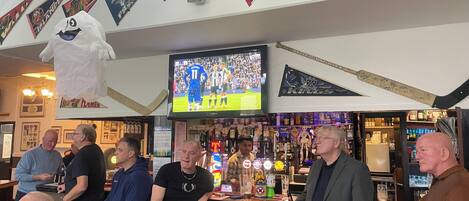 Un bar con una televisión mostrando un evento deportivo, varias banderas y un ventilador de techo.