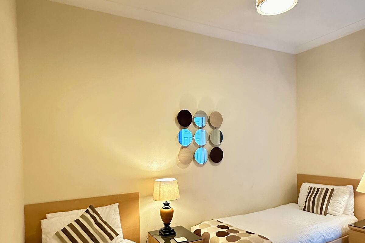 A hotel room with two beds, a nightstand with a lamp, a wall-mounted mirror, and a ceiling light fixture.