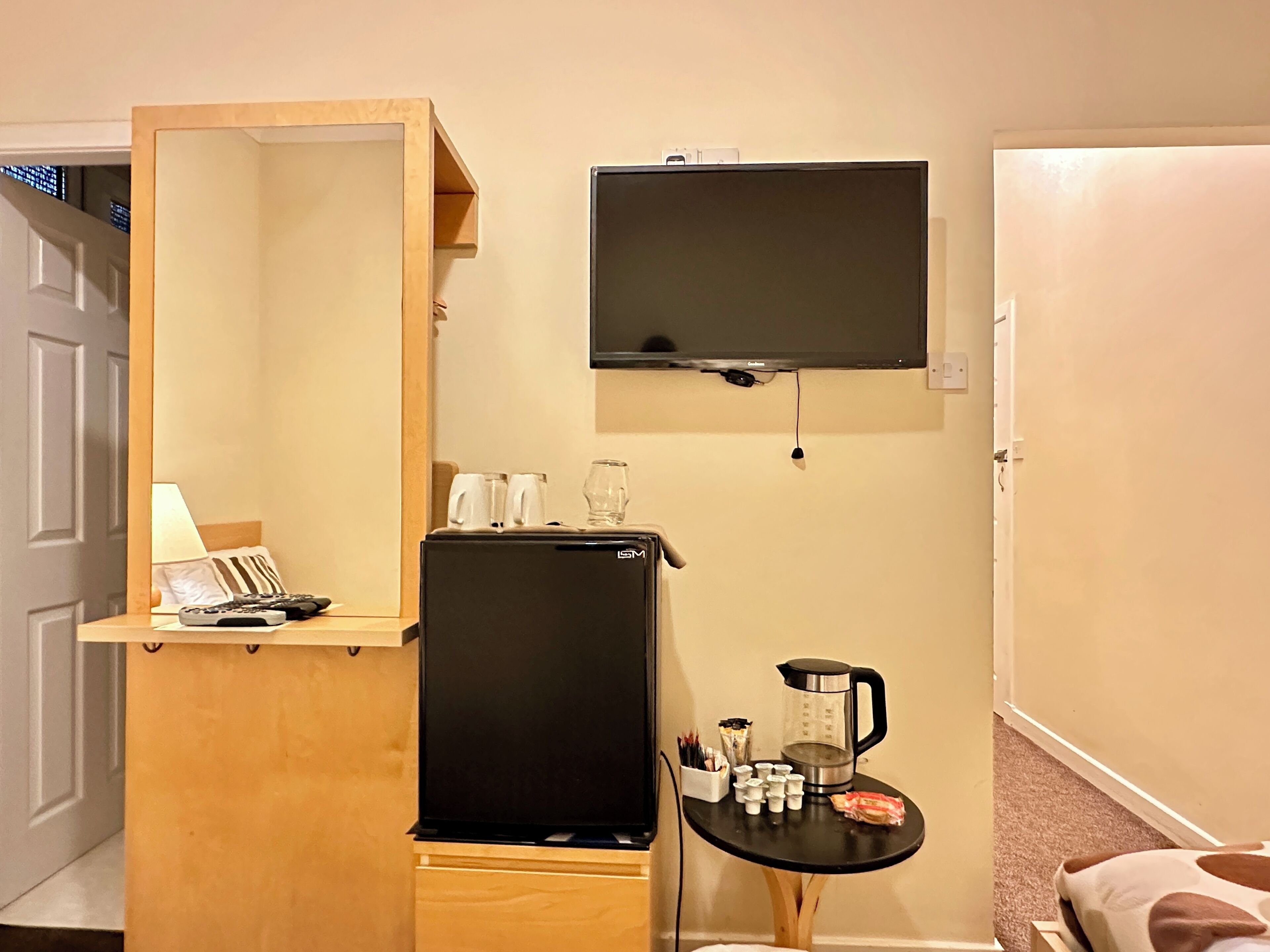 Een compacte hotelkamer met een houten kast, een zwarte koelkast, een klein tafeltje met een waterkoker, kopjes en een zakje snacks. Een televisie is aan de muur boven de kast bevestigd.