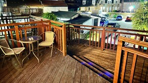 A wooden deck with two wicker chairs and a small table, overlooking a street with parked cars and buildings. - The Elbow Room (Kirkcaldy)