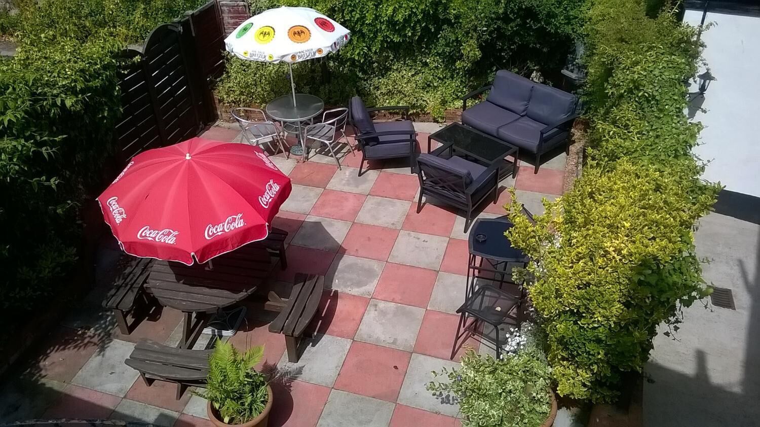 Un patio al aire libre con un paraguas rojo de Coca-Cola, una mesa redonda y sillones de descanso negros.