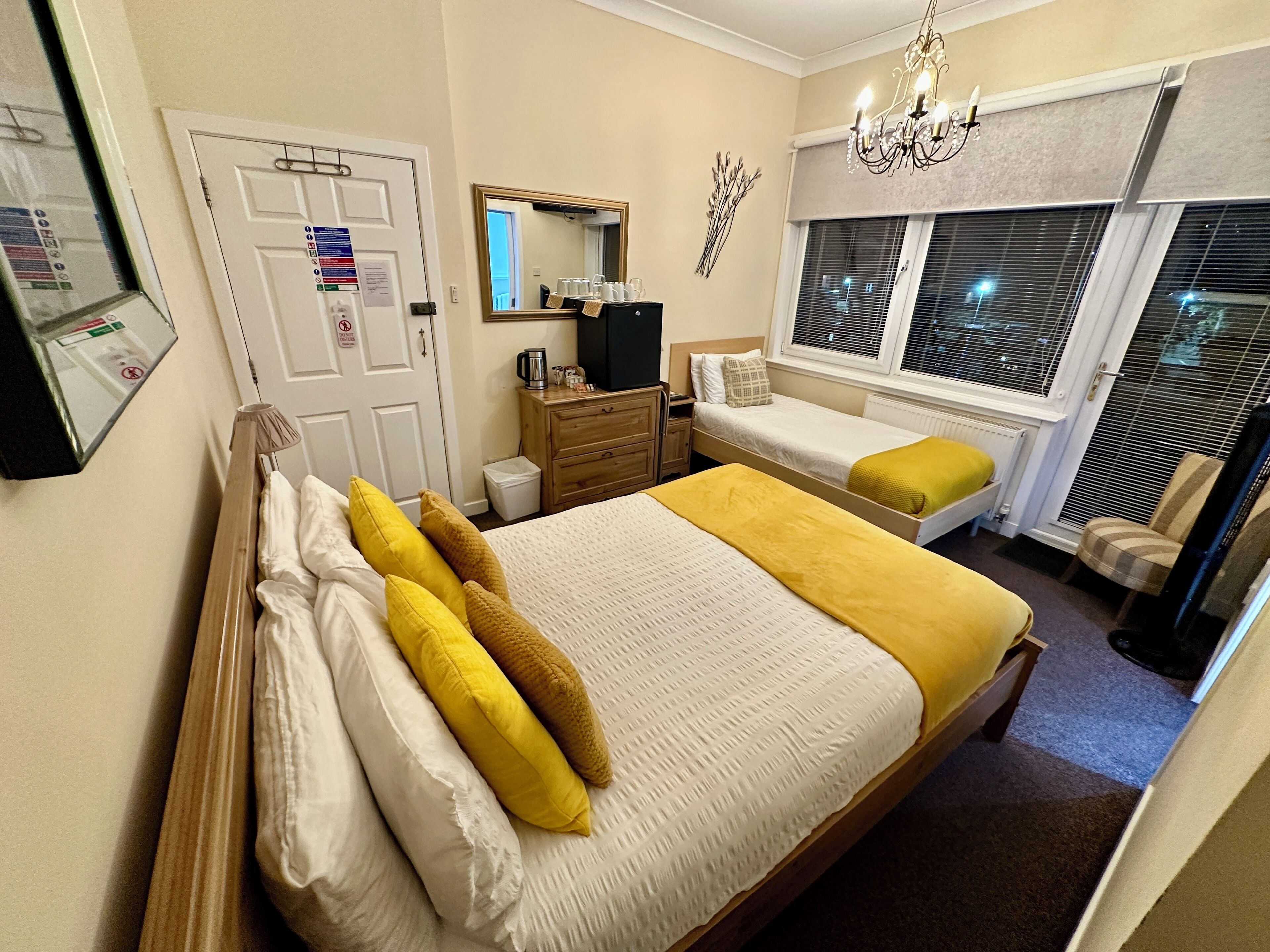 Une chambre à coucher avec un grand lit, une commode, un miroir, une fenêtre avec des stores et un lustre.