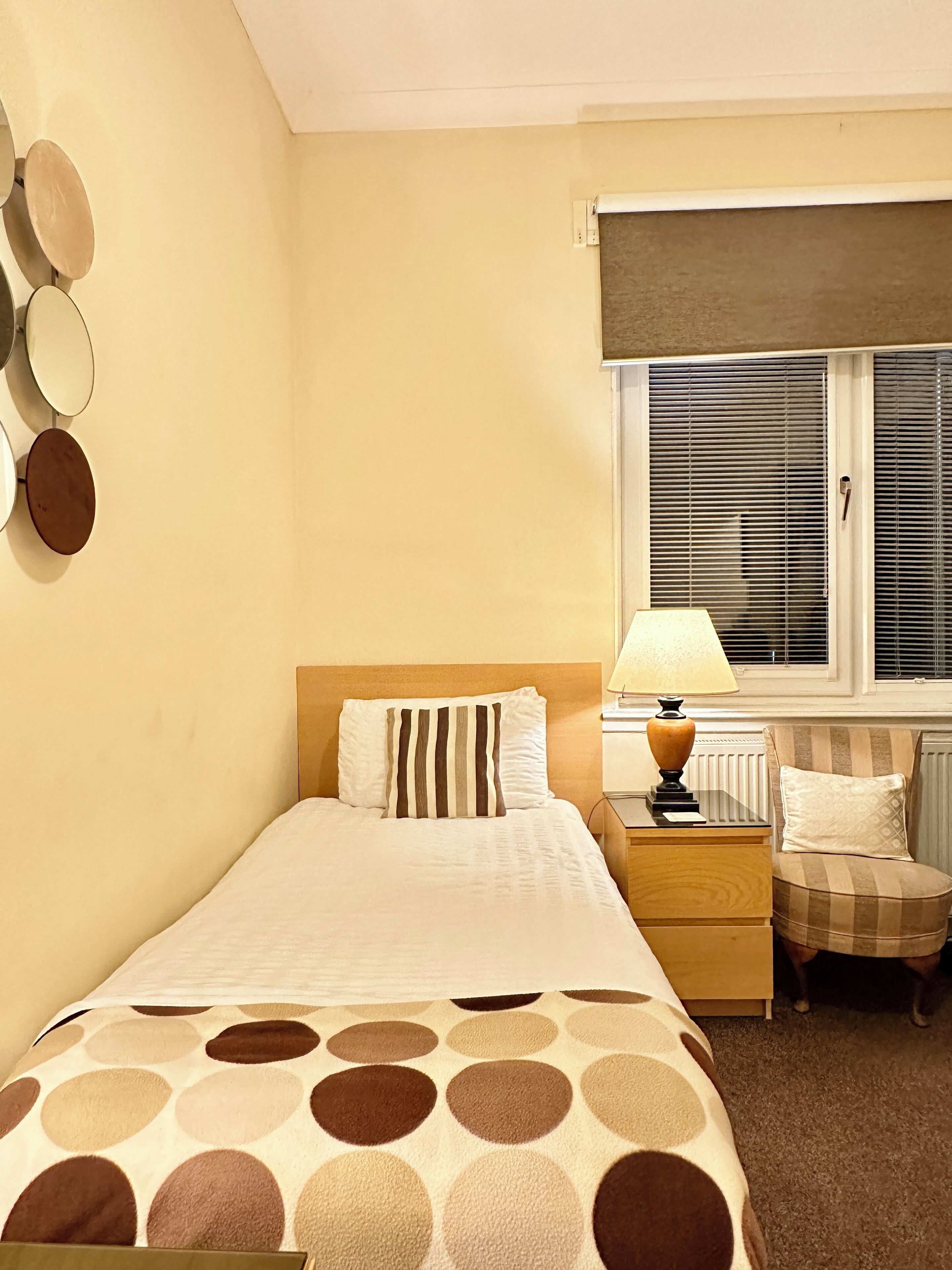 A hotel room with a bed, a nightstand, a lamp, a chair, and a window with blinds.