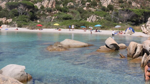 Ein Strand mit klarem Wasser, Felsen und Menschen.