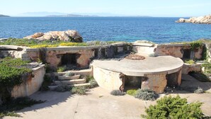 Ein Küstenbereich mit Außenbestuhlung, Stein-Tische und -Stühle, umgeben von Vegetation mit Blick aufs Meer.