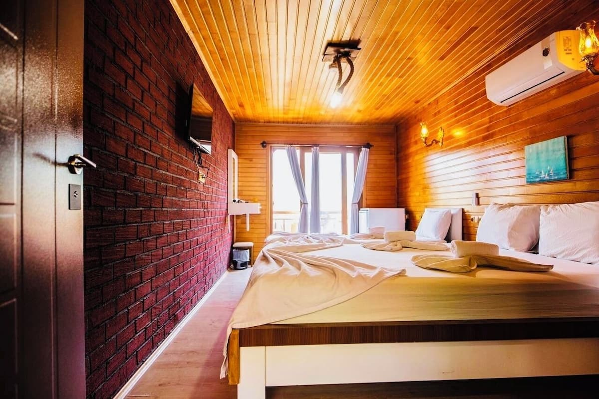 Una habitación con pared de ladrillo, techo de madera, una cama con ropa blanca y una ventana con cortinas.