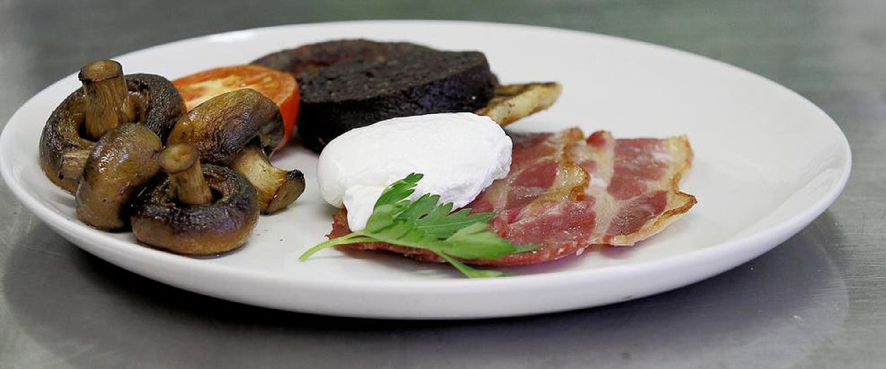 Un plato con champiñones, una rodaja de tomate, una loncha de morcilla, un huevo escalfado y una rebanada de tocino.