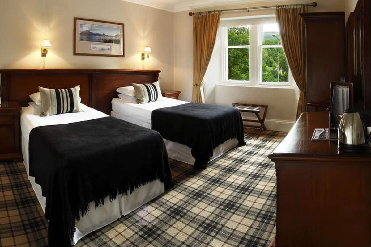 A hotel room with two beds, a wooden headboard, a plaid floor, a window with curtains, and a kettle on the desk.