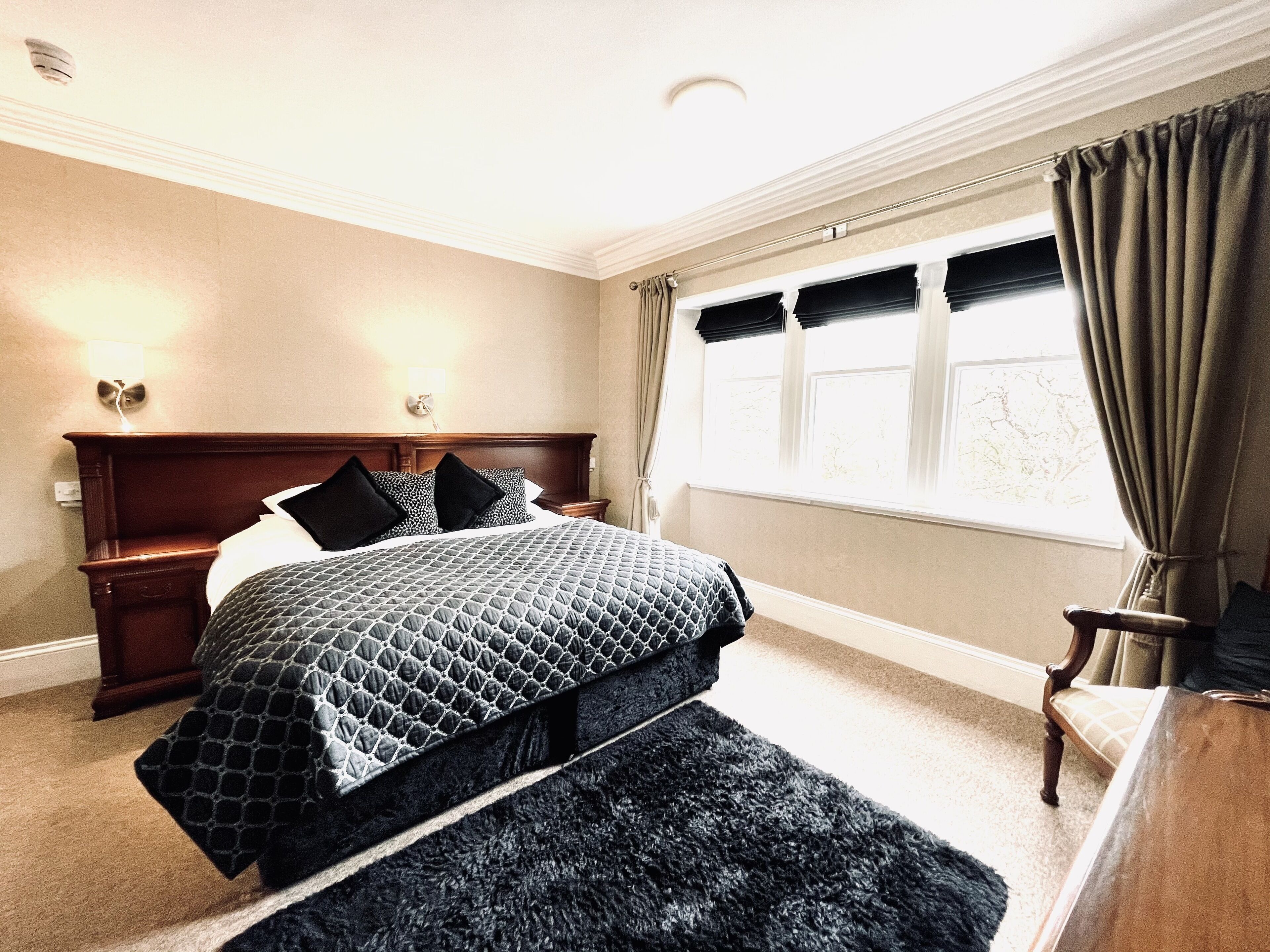 Una habitación de hotel con una cama grande, una mesita de noche, un aparador de madera y una ventana con cortinas.