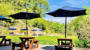 An outdoor seating area with wooden tables and blue umbrellas, surrounded by lush greenery and trees. - Invergarry Hotel (Invergarry)