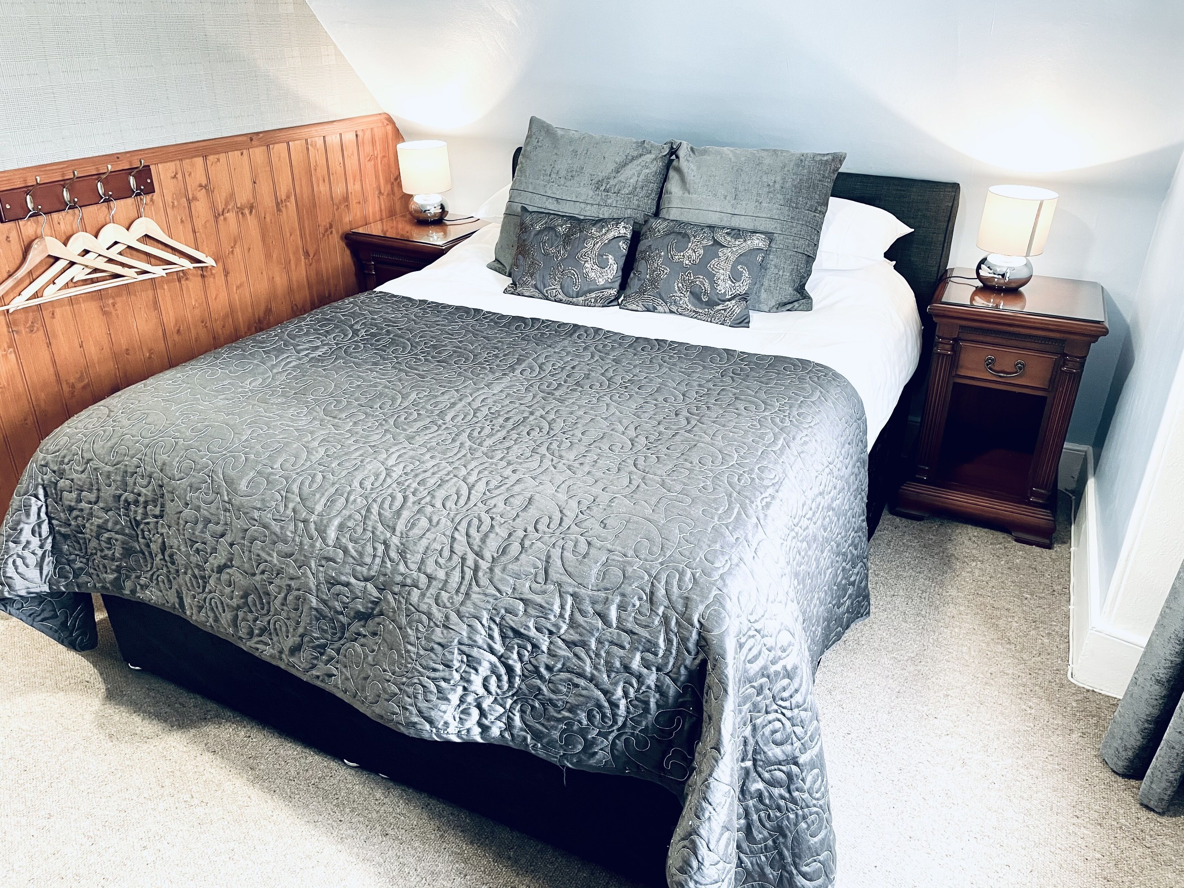 Una cama bien tendida con una colcha estampada, cabecera de madera, dos mesitas de noche con lámparas y un perchero de madera.