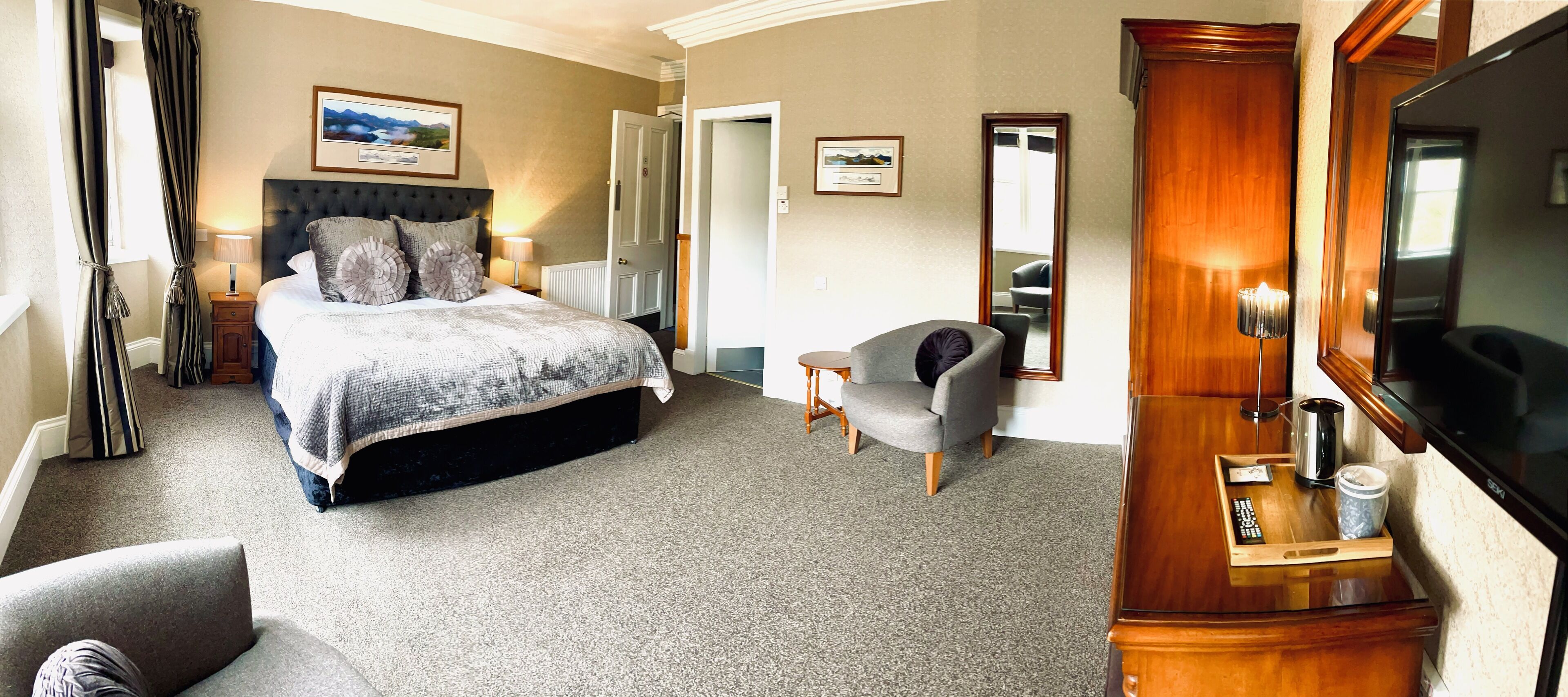 A hotel room with a large bed, a grey armchair, a wooden TV stand, and a mirror.