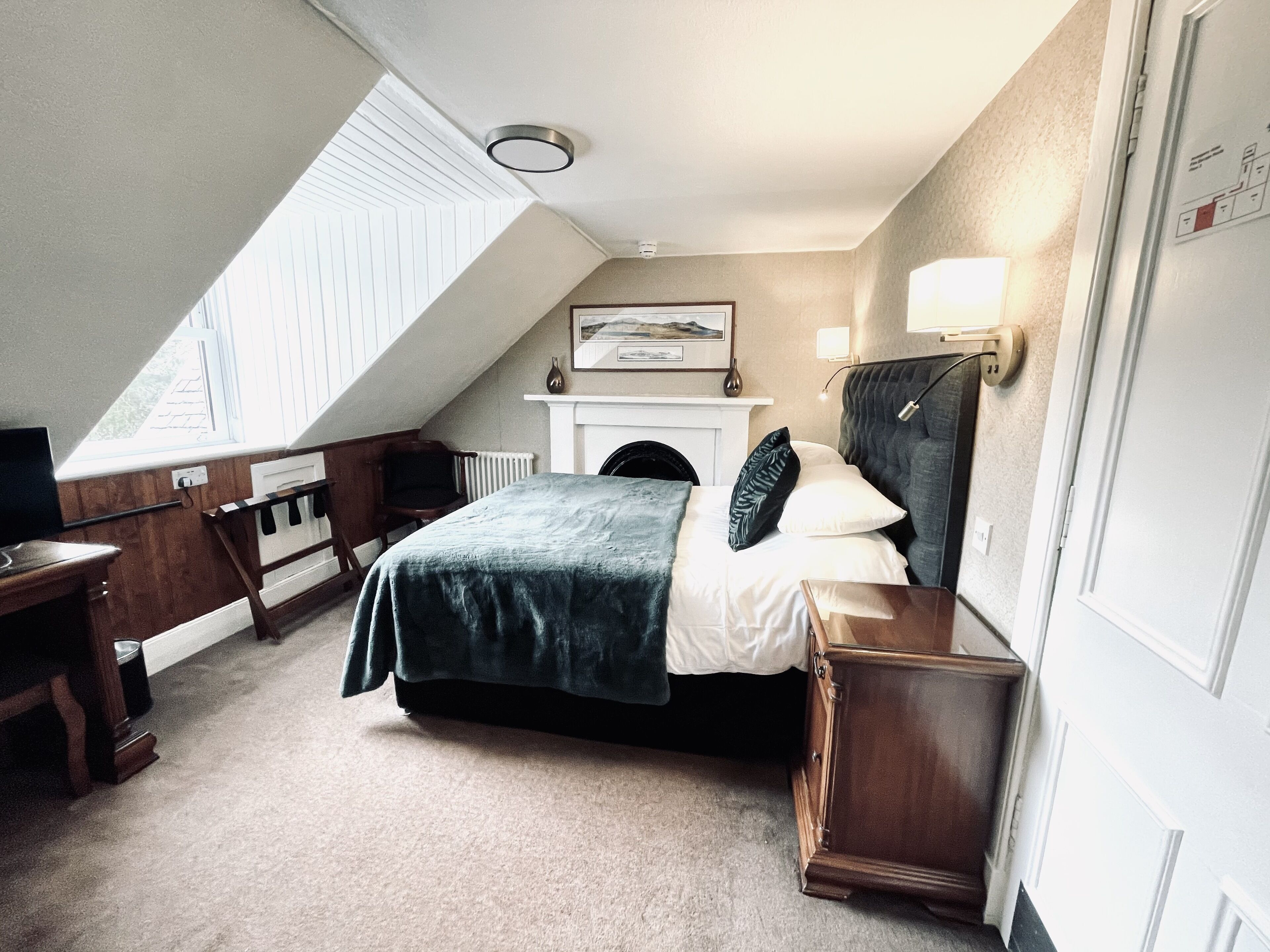 A bedroom with a sloped ceiling, a bed with a dark green bedspread, a bedside table, a lamp, and a fireplace.