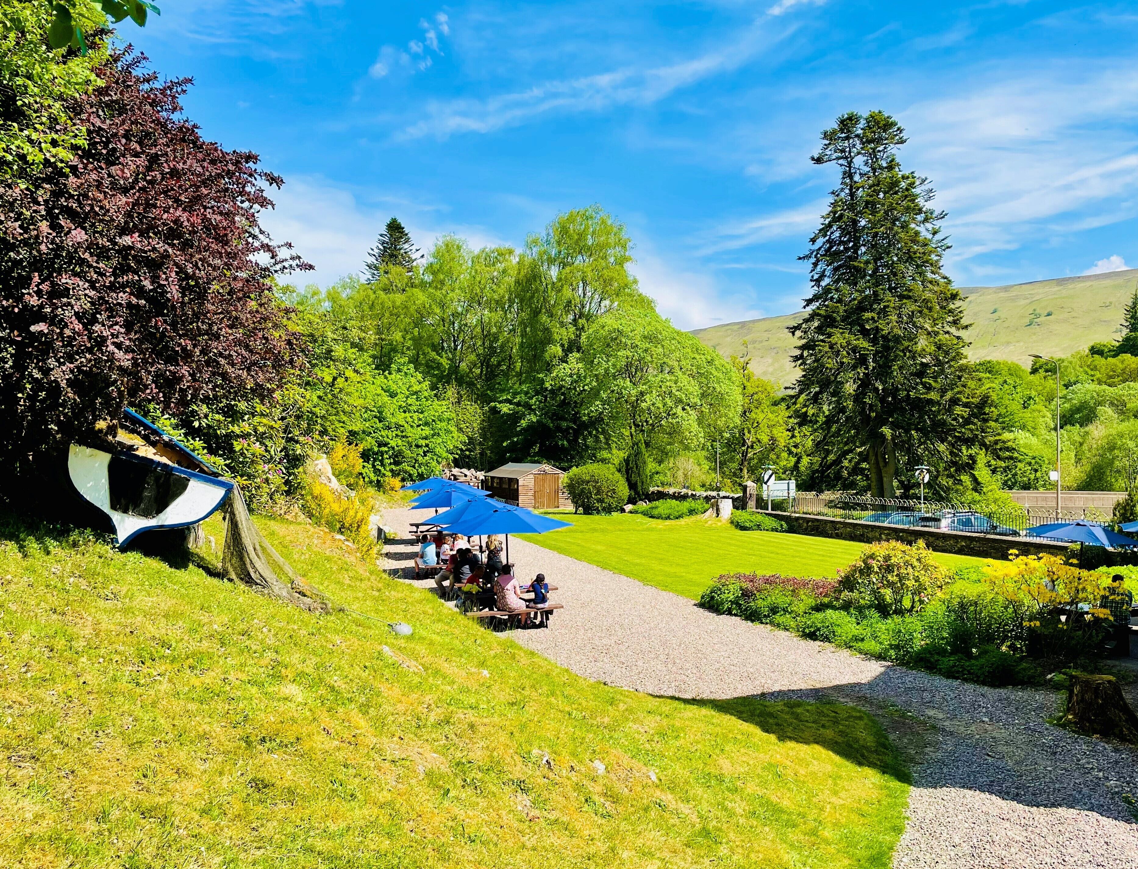 A picnic area with tables and blue umbrellas, surrounded by lush greenery and trees.