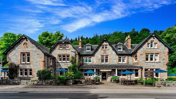 A stone building with multiple windows and a central entrance, surrounded by greenery and outdoor seating. - Invergarry Hotel (Invergarry)