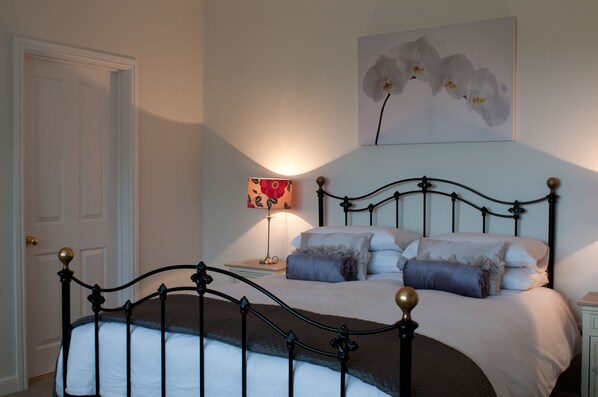 A neatly made bed with a black metal frame and white bedding, a bedside table with a lamp, and a door to the left.