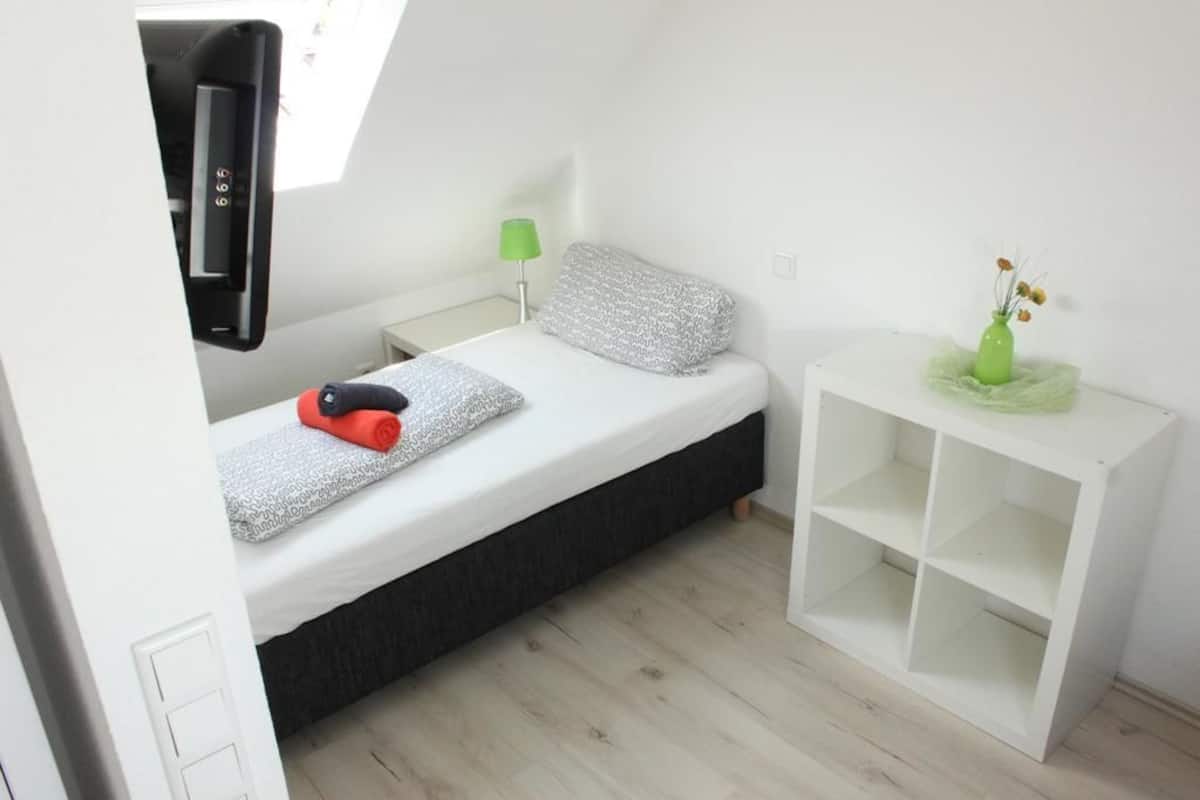 Un dormitorio pequeño y ordenado con cama, mesita de noche, lámpara y estantería blanca.