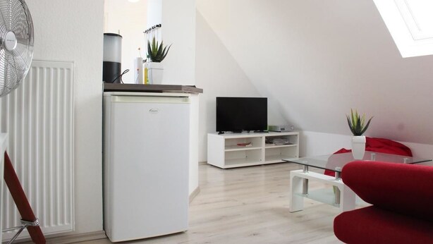 A modern living room with a white refrigerator, a flat-screen TV on a white stand, a red sofa, and a small table. - Ferienwohnung Fulda (Fulda)