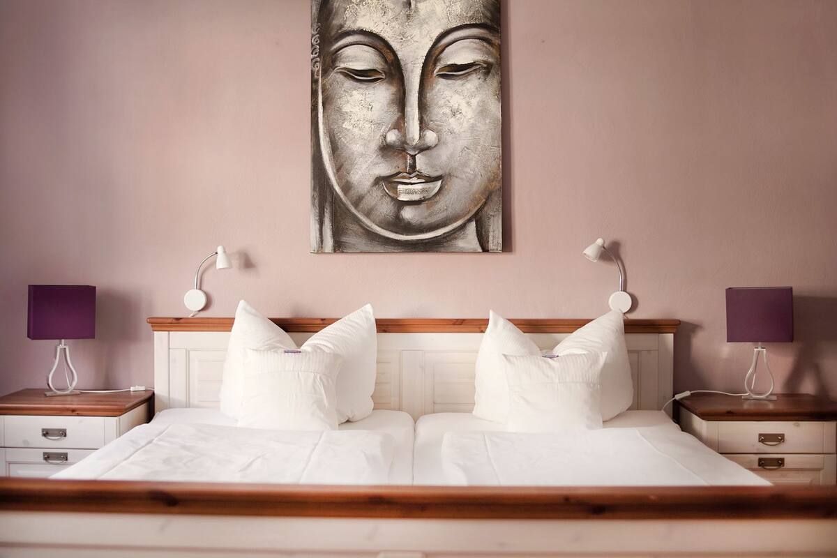 Ein Schlafzimmer mit einem Holzkopfteil, weißer Bettwäsche, zwei Nachttischen mit Lampen und einem großen, abstrakten Gemälde an der Wand.