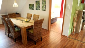 A dining area with a wooden table and wicker chairs, a hanging light fixture, and a view into another room with a sofa and a red curtain. - Haus Am Fluss (Laurenburg)