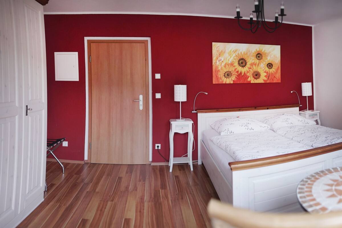 Ein Schlafzimmer mit Holzboden, einem Bett mit weißem Kopfteil, einem runden Tisch mit gemustertem Tischläufer, einer roten Wand mit einem Sonnenblumenbild und einer Chandelier.