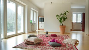 Un salon spacieux avec de grandes fenêtres, une plante en pot, un tapis aux motifs rouges et blancs, et une petite table sur laquelle est posée une lanterne.