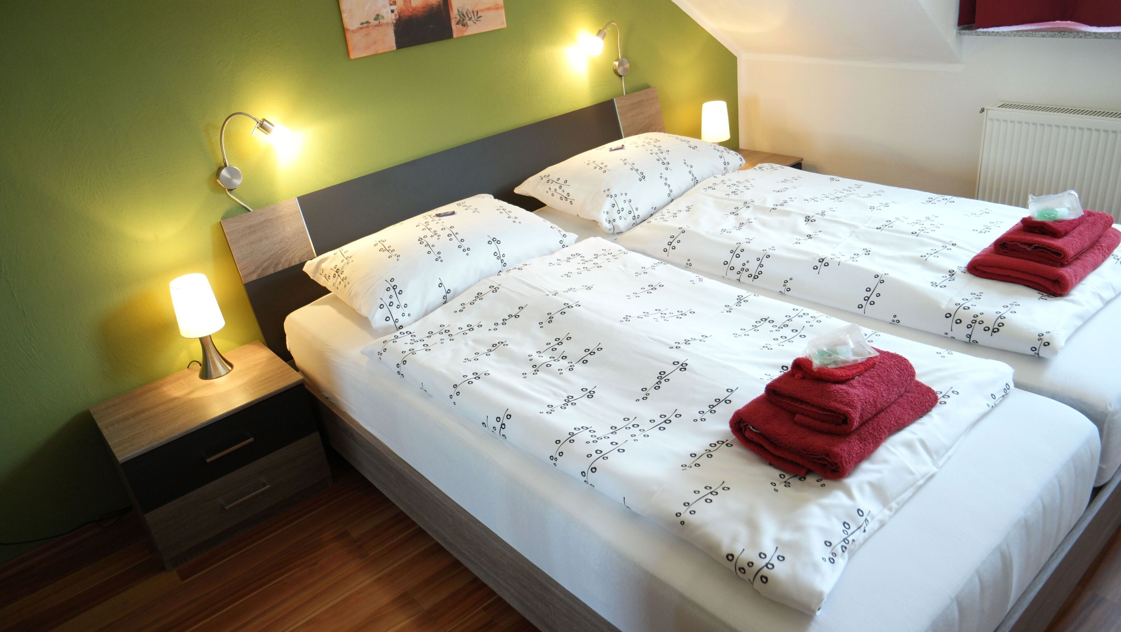 A twin room with two beds, each with white bedding and a floral pattern, two bedside lamps, and a small wooden nightstand with a drawer.