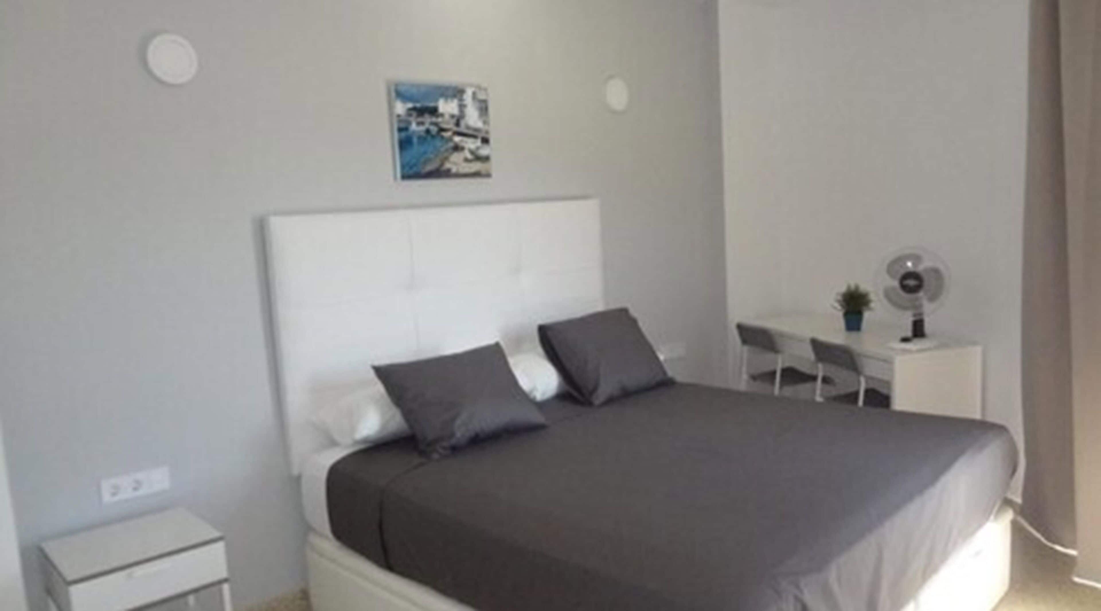 Une chambre à coucher avec un grand lit, un petit bureau, une chaise et un ventilateur fixé au mur.