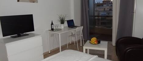A hotel room with a bed, a desk with a laptop, a television, a small table with fruit, and a balcony with a view of buildings.