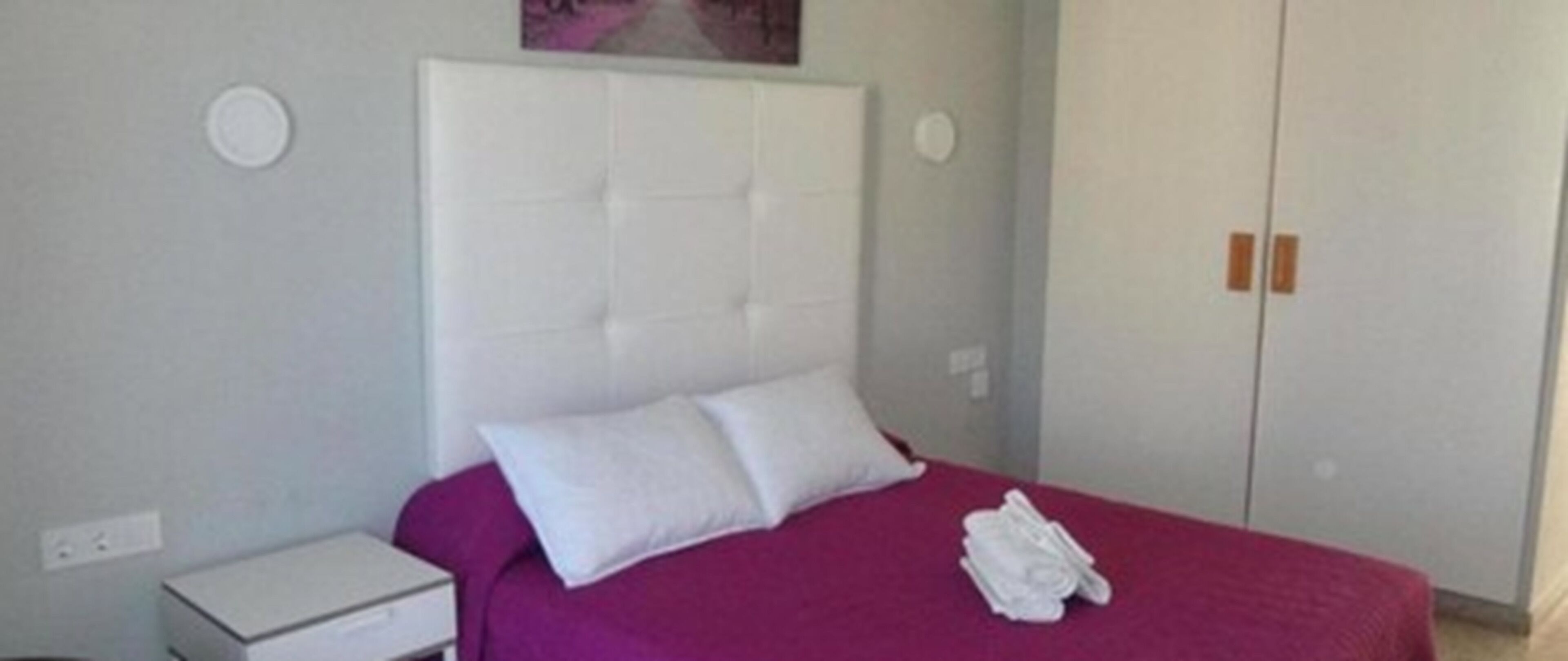 Een slaapkamer met een bed, een wit hoofdeinde en een paarse plaid. Er staat een witte nachtkastje en een witte garderobe met twee deuren.