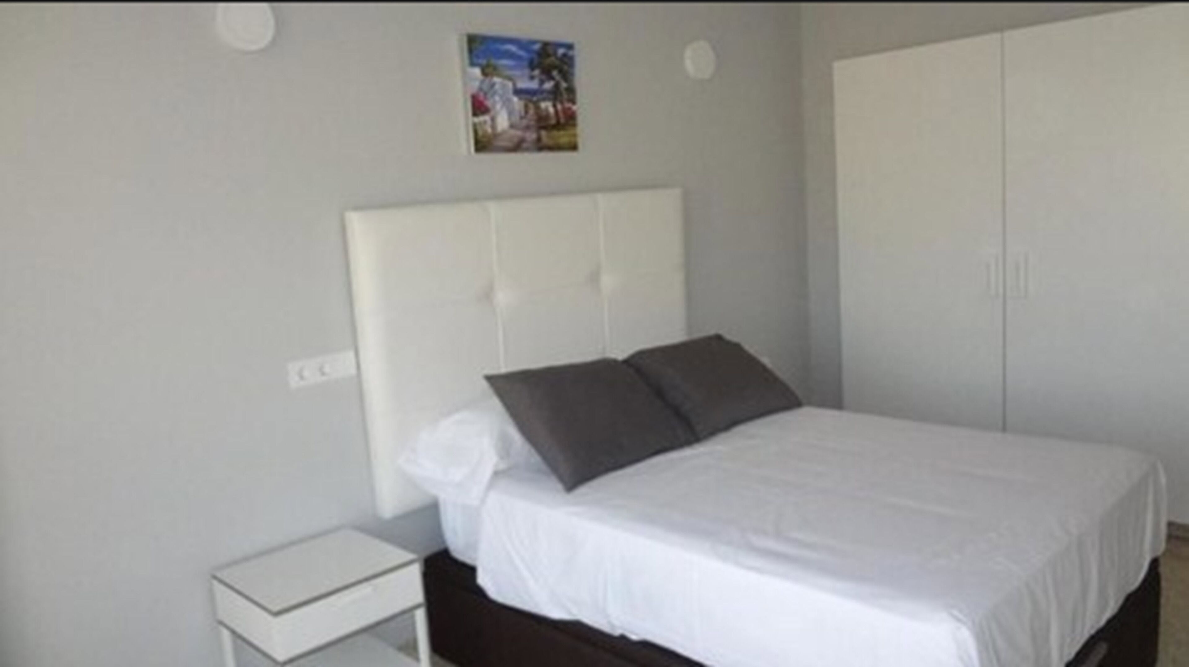A bedroom with a white bed, two pillows, a nightstand, and a wardrobe.
