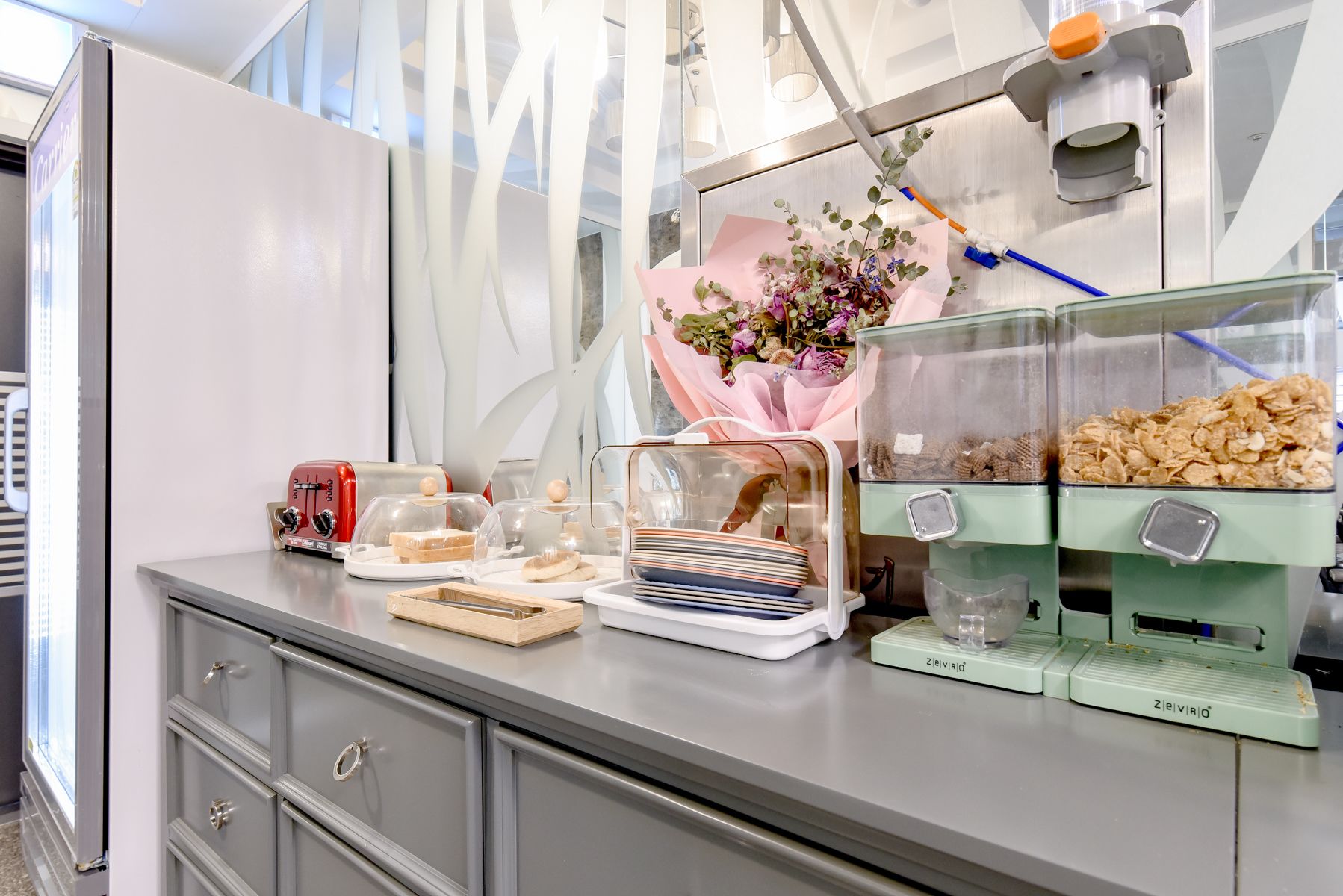 A buffet setup with a toaster, a glass display case with food, and a flower arrangement.