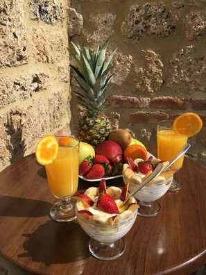 A table with a fruit bowl, two glasses of orange juice, a pineapple, and a variety of fruits. - Varosi 4 Seasons (Edessa)