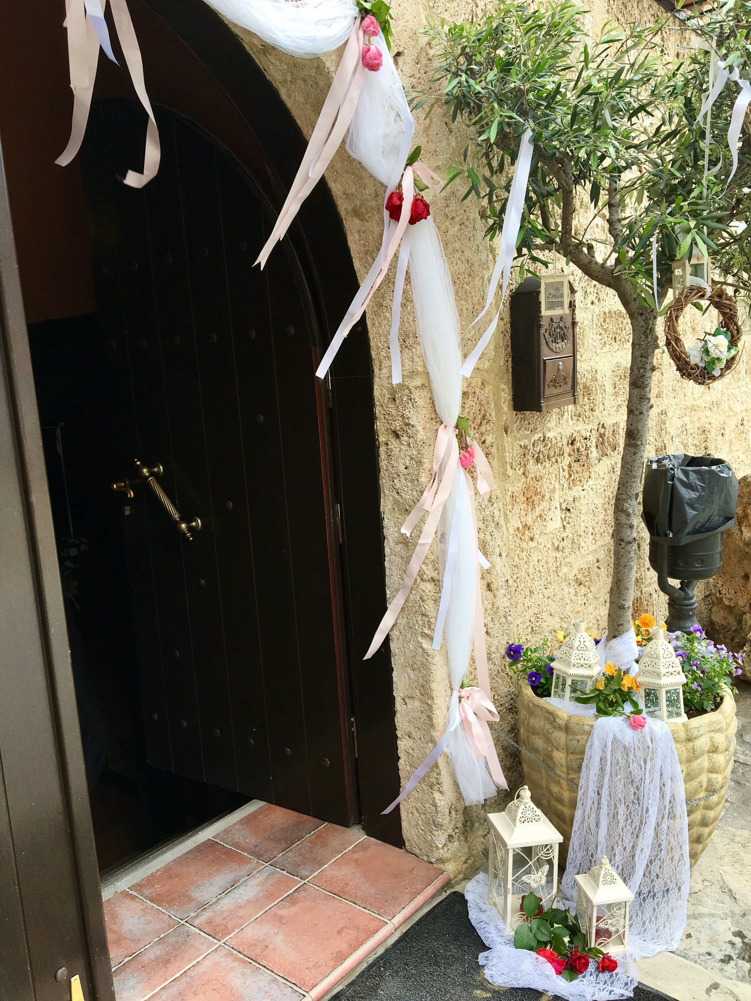 A stone building entrance with a dark wooden door, a potted plant with flowers, and a small white lantern.