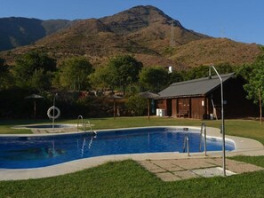 Una piscina con montañas al fondo, un edificio de madera y una zona con césped.