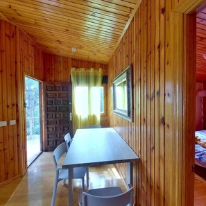 Interior de una cabaña de madera con una mesa de comedor y sillas, una pared de piedra y una ventana con cortinas.