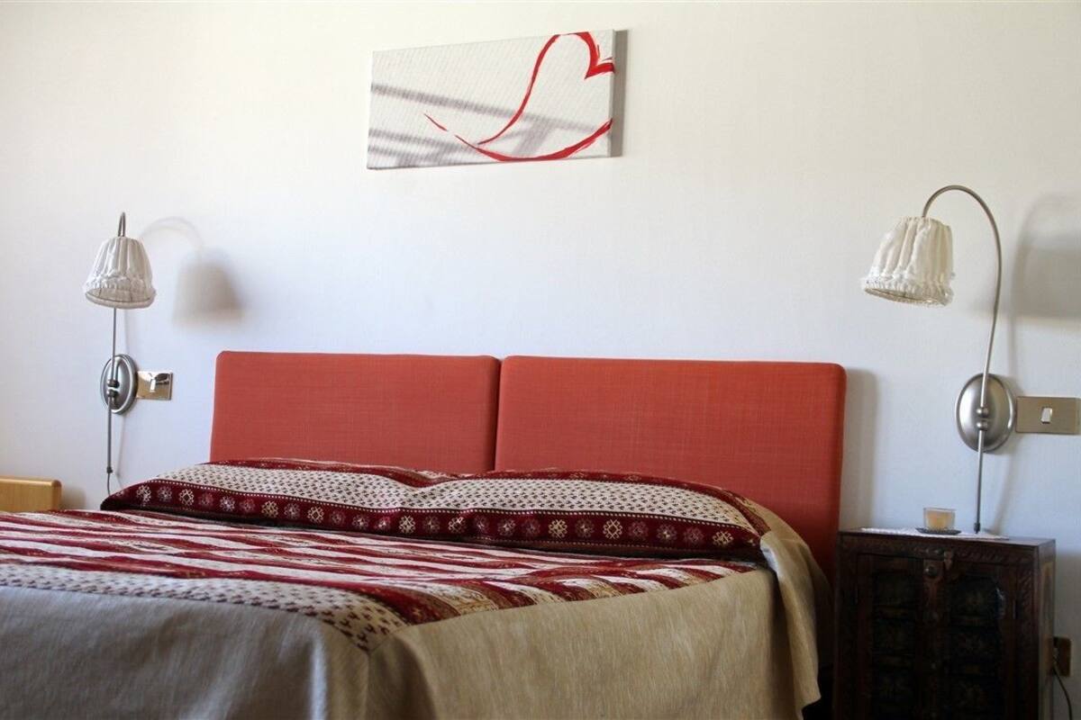 A bedroom with a red headboard, a patterned bedspread, two bedside lamps, and a wall-mounted abstract art piece.