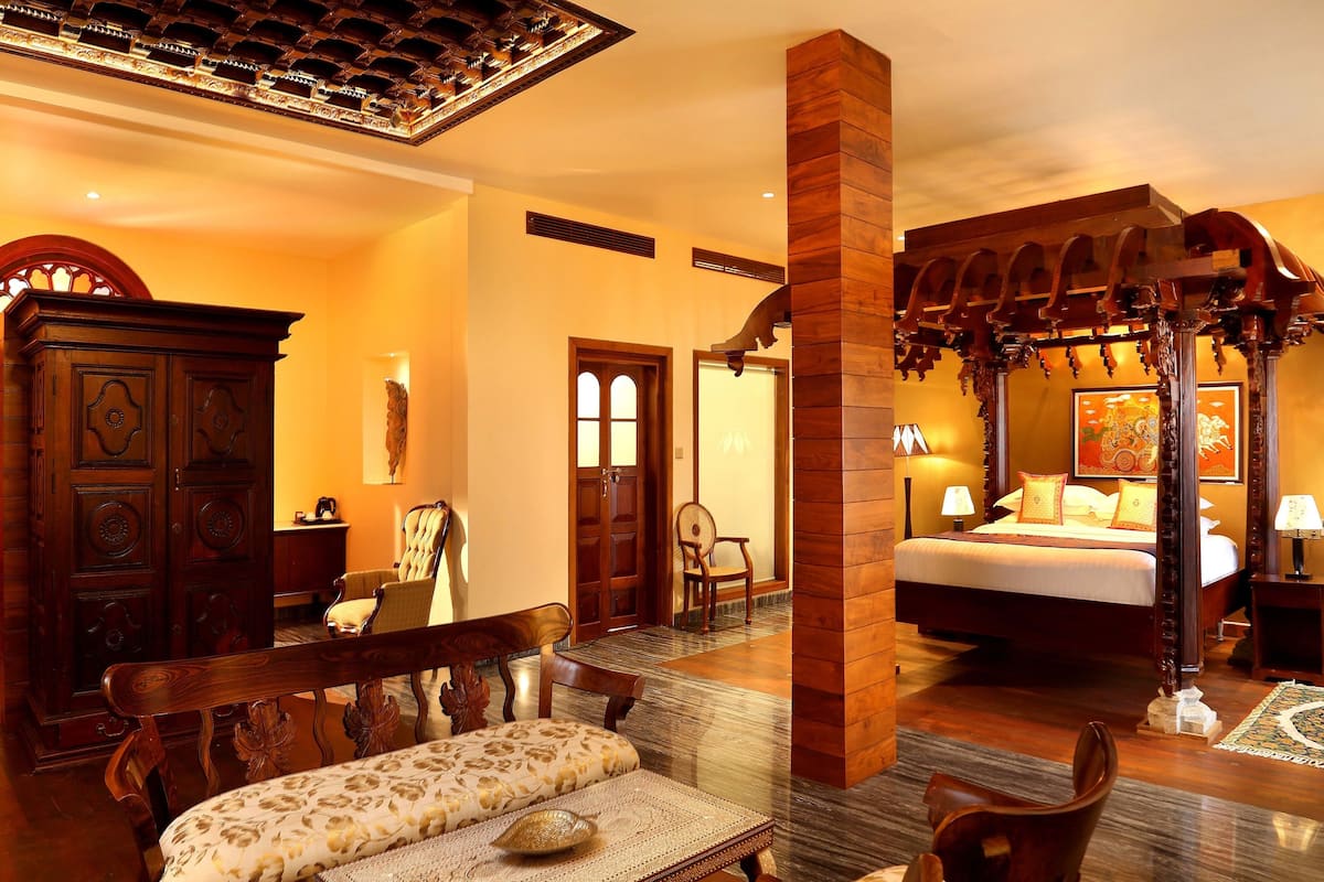 A spacious hotel room with a large bed, a sitting area with a bench, a wooden wardrobe, and a dining area with a table and chairs.