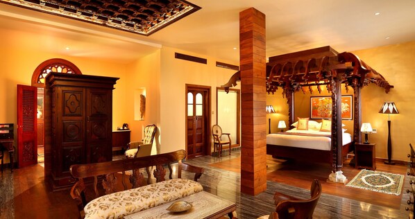 A spacious hotel room with a large bed, a sitting area with a bench, a wooden wardrobe, and a dining area with a table and chairs.